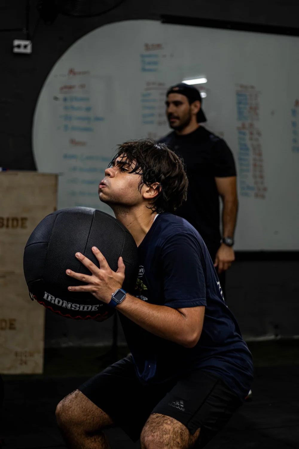 Participante haciendo sentadillas con balón medicinal acompañado por un entrenador